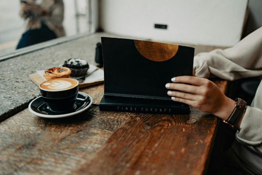 Personne lisant un livre avec un café et des pâtisseries sur une table en bois, près d’une fenêtre dans un café cosy.