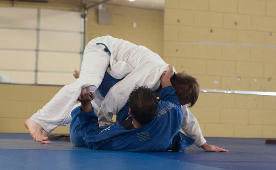Two practitioners in blue and white gis engaged in a Brazilian Jiu-Jitsu sparring session, one executing a sweep from the bottom position.