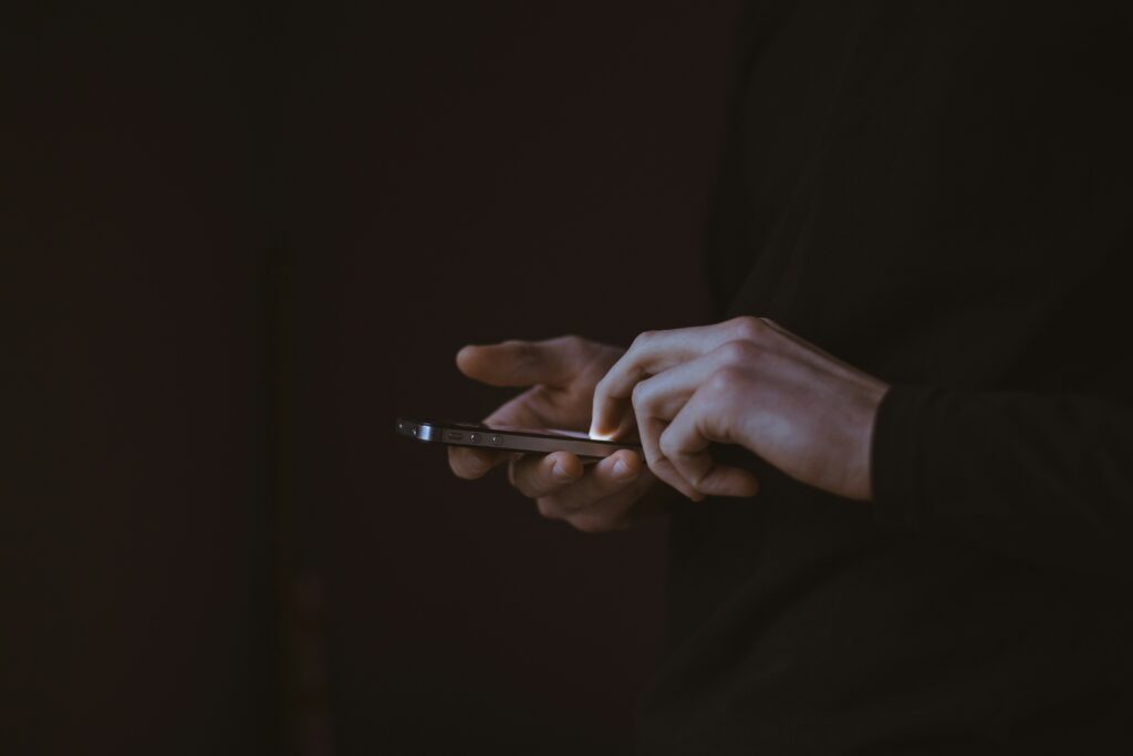 Mains d’une personne utilisant un smartphone dans une pièce sombre, lumière de l’écran visible.